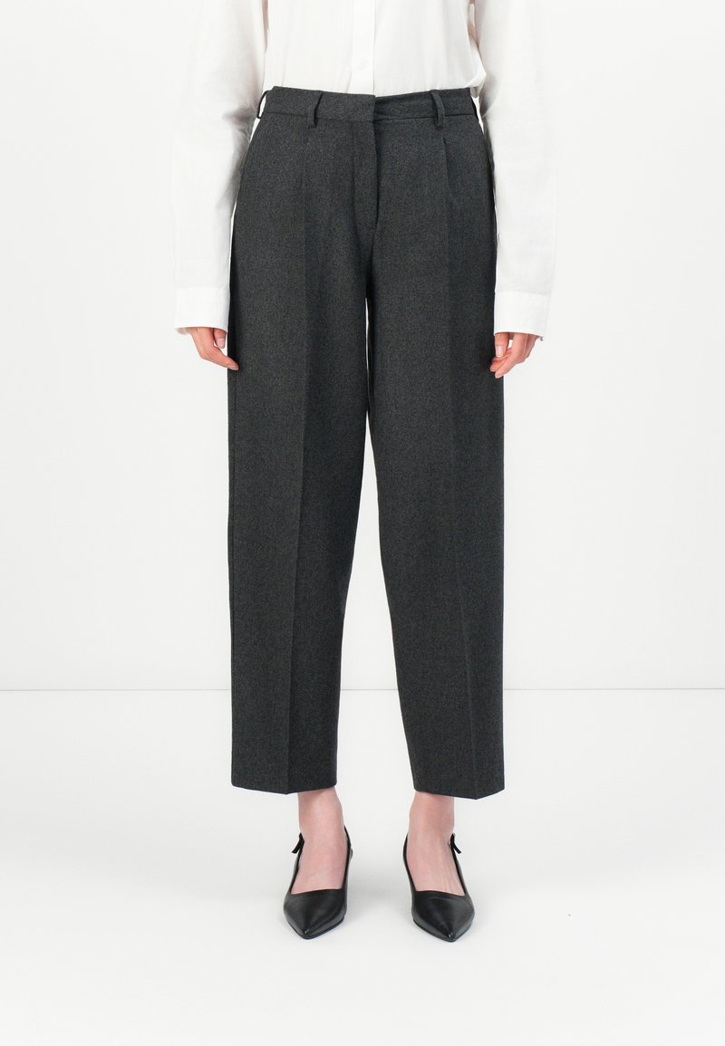 Personne portant un pantalon tailleur gris foncé cropped, une chemise blanche à manches longues et des escarpins noirs à bout pointu, se tenant sur un fond blanc.