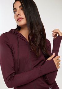 Donna con lunghi capelli scuri che indossa una felpa rossa con cerniera e aperture per i pollici, in posa con il braccio sinistro incrociato e guardando di lato.