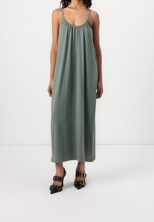 Femme portant une robe longue verte ample sans manches avec des fines bretelles et des talons noirs à bouts pointus avec des larges brides et des boucles.