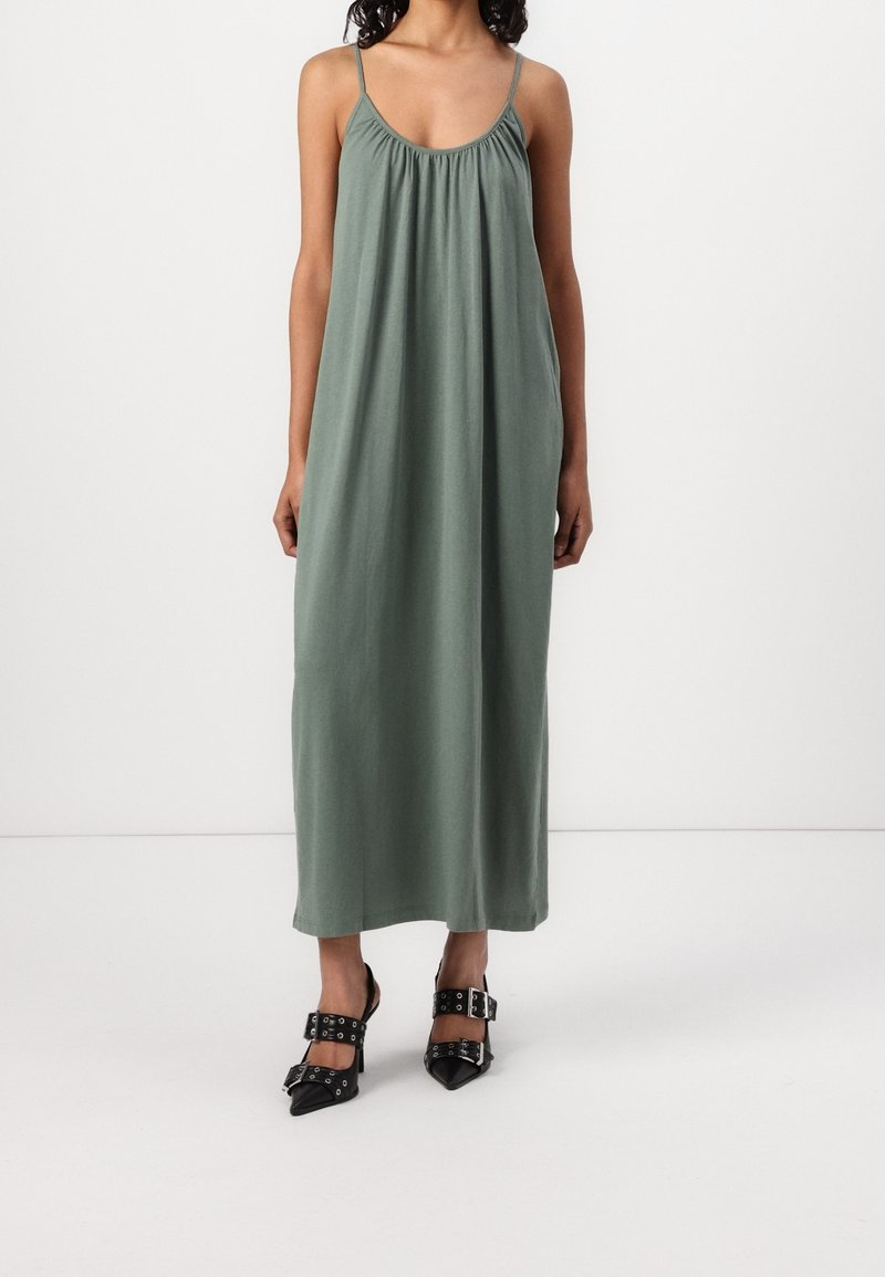 Femme portant une robe longue verte ample sans manches avec des fines bretelles et des talons noirs à bouts pointus avec des larges brides et des boucles.