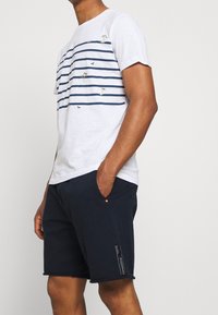 Weißes T-Shirt mit kurzen Ärmeln und navyblauen horizontalen Streifen, mit Palmen- und Vogelmotiven. Kombiniert mit einfarbigen navyblauen Shorts.
