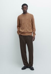 Brauner Strickpullover mit Rundhalsausschnitt und gerippten Bündchen, kombiniert mit locker sitzender brauner Hose und schwarzen Lederschuhen. Minimalistischer Hintergrund.