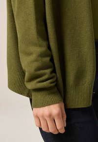 Olivově zelený pletený cardigan s žebrovanými manžetami, měkkou texturou a volným střihem, zobrazený v detailním záběru rukávu a ruky.