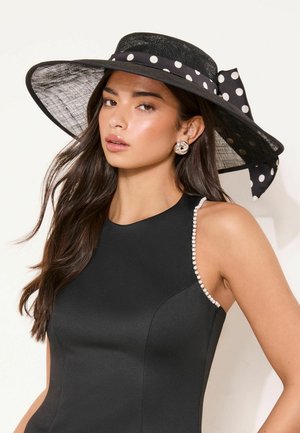 Woman wearing a black sleeveless dress with pearl trim and a wide-brim black hat with a white polka dot ribbon bow.