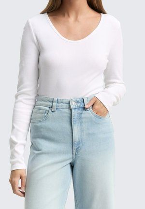 Frau trägt ein weißes Langarmshirt mit Wasserfallausschnitt, in hochtaillierte hellblaue Jeans gesteckt, mit einer Hand in der Tasche.