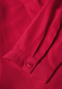 Détail d'une chemise rouge avec un tissu texturé, fermeture à bouton sur le poignet, et un tombé doux avec une finition légèrement brillante.