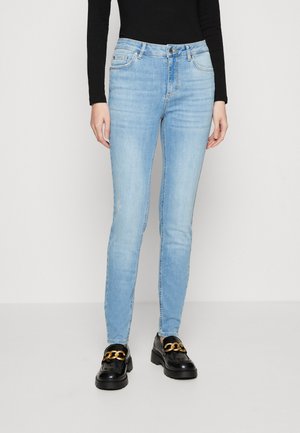 Vaqueros skinnies de color azul claro en material de denim con bolsillos frontales y cierre de botón, combinados con mocasines negros que tienen un acento de cadena dorada.