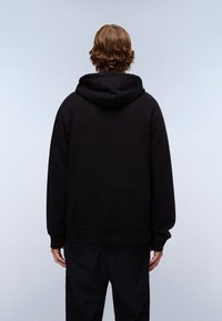 Persona con capelli castani ondulati che indossa una felpa nera con cappuccio e pantaloni neri, in piedi di spalle contro uno sfondo chiaro e uniforme.