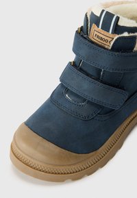 Zili sintētiski potīšu zābaki ar bežīgu vilnas oderi, diviem Velcro siksnām un strukturētu brūnu gumijas zoli. Iekļauj kontrastējošu šuvumu un logo birku.