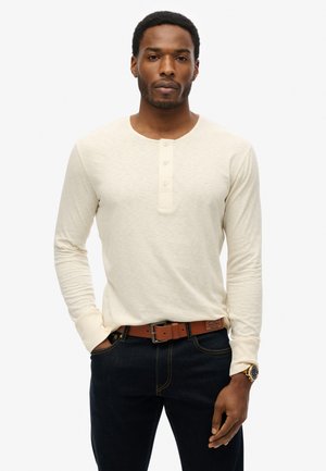 Lange mouw crèmekleurige henley shirt met drie knopen bij de halslijn, gecombineerd met donkere jeans en een leren bruine riem. Glad stoftextuur.