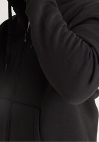 Zwarte zip-up hoodie gemaakt van zachte, gestructureerde stof. Voorzien van een zak aan de voorkant en een trekkoord. Close-up van de mouw en de rits.