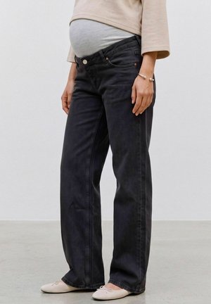 Personne enceinte portant un jean de maternité noir ample par-dessus une ceinture de ventre grise avec un haut beige et des ballerines blanches.