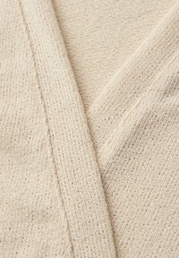 Zbliżenie kremowej dzianiny z widocznym fakturowanym splotem i nakładającymi się krawędziami tworzącymi wzór w kształcie litery V.