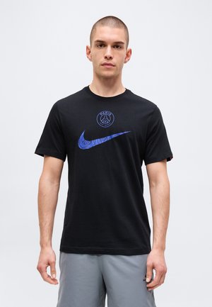 Młody mężczyzna z krótkimi włosami, ubrany w czarną koszulkę z niebieskimi logo Paris Saint-Germain i Nike oraz jasnoszare szorty, stoi na jednolitym tle.