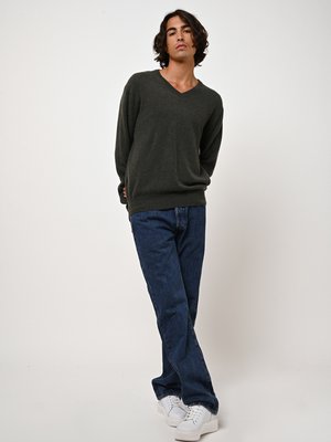 Grüner V-Ausschnitt-Pullover aus weichem Stoff, kombiniert mit dunkelblauen Jeans und weißen Sneakers. Das Modell posiert in einer entspannten Haltung.
