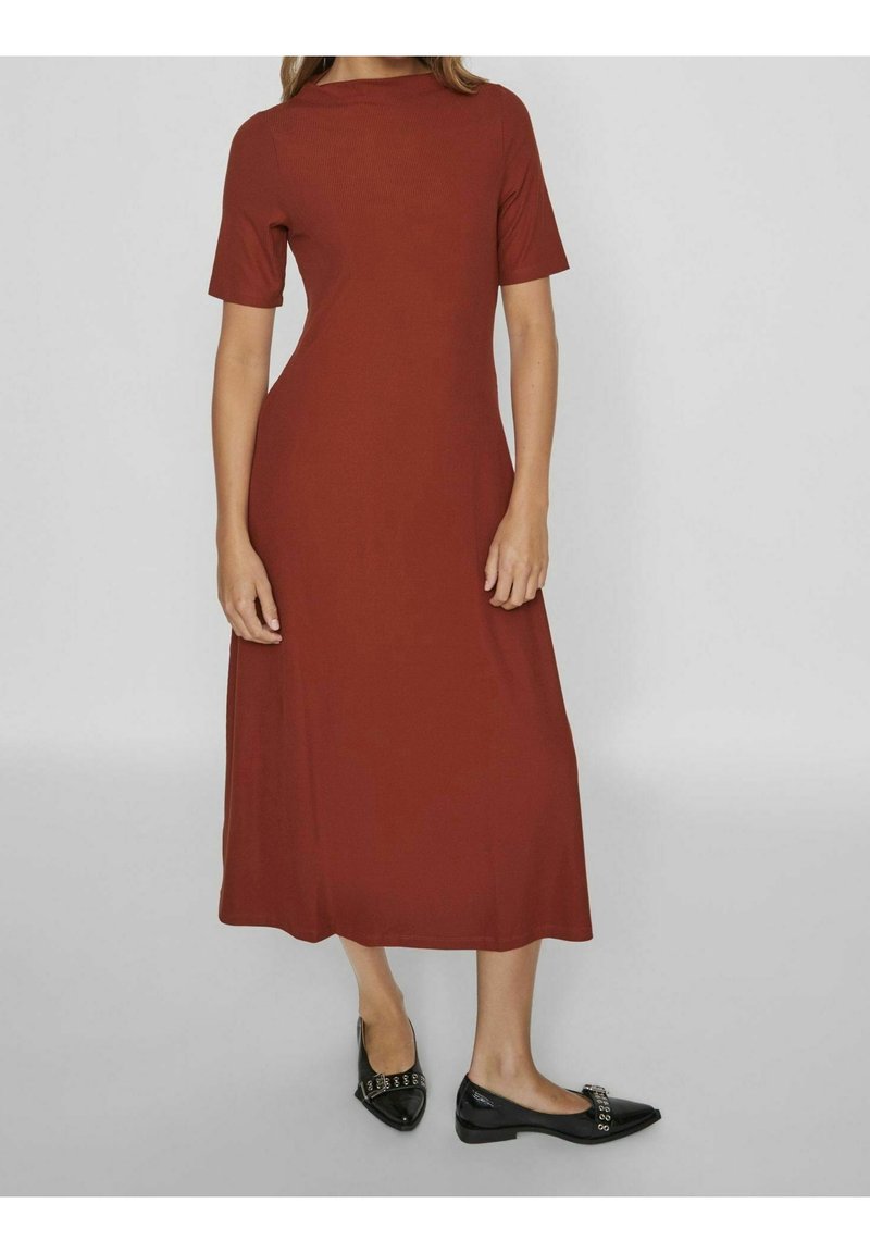 Femme portant une robe midi rouille à manches courtes associée à des ballerines noires pointues avec des clous argentés, debout devant un fond gris uni.