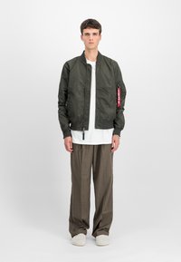 Jachetă bomber verde măsliniu cu fermoar frontal, manșete cu nervuri și un etichetă decorativă roșie. Asortată cu pantaloni largi gri deschis și o cămașă albă.