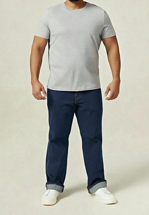 Homme portant un t-shirt gris clair à manches courtes, un pantalon bleu foncé retroussé et des chaussures blanches sans lacets, debout devant un fond uni.