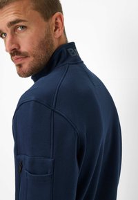 Navy blue fleece jacket with a high collar, side zip pocket, and textured shoulder seams. Features a logo on the collar.