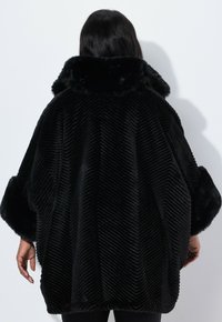 Poncho in velluto a coste nero con colletto foderato in pelliccia, caratterizzato da un motivo a spina di pesce testurizzato e un taglio morbido e oversize, visto da dietro.