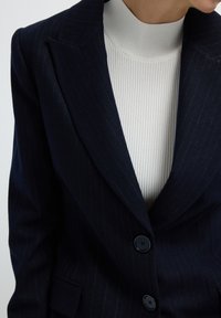 Námořnický pruhovaný blazer s širokými klopami, třemi knoflíky a jednou náprsní kapsou, vrstvený přes bílý žebrovaný rolák.