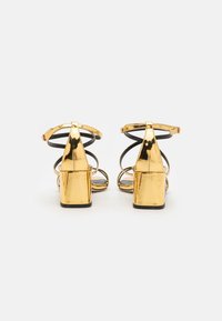 Gold heeled sandals featuring a shiny patent finish, open toe, and black ankle straps with a block heel design.