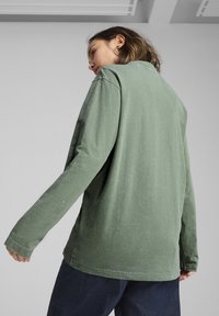 Cămașă cu mânecă lungă din bumbac verde estompat, cu o croială lejeră, având guler rotund și umeri ușor lăsați. Textură moale, detalii minimale.