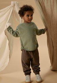 Jeune enfant aux cheveux bouclés portant un pull vert et un pantalon marron, tenant des rideaux beige et marron clair dans un décor aux tons neutres.