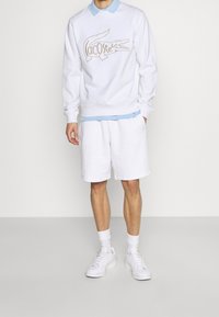 Sweatshirt blanc avec logo Lacoste beige, associé à une chemise à col bleu clair, un short blanc et des baskets blanches, sur un fond neutre.