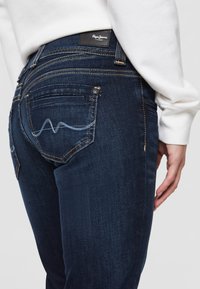 Tmavě modré džíny z denimu s přiléhavým designem. Mezi funkce patří kontrastní stehy, malá nášivka na pase a zakřivené zadní kapsy.