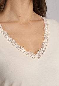 Top beige à col en V avec une délicate bordure en dentelle le long de l'encolure, texture lisse et design ajusté.