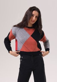 Pull en tricot avec un motif en diamant rouge, gris et noir. Design court avec des poignets et un ourlet côtelés, détails en fil texturé.