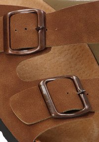 Dos correas de cuero marrón con hebillas de metal rectangulares sobre una superficie texturizada color beige, mostrando agujeros de ajuste.