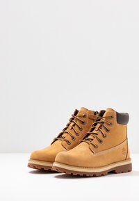 Bottes de cheville en suede beige avec des accents en cuir marron, œillets en métal et semelles en caoutchouc beige ; présente des détails cousus et un logo sur le côté.