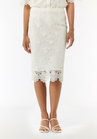 Jupe en dentelle blanche avec un ourlet festonné, présentant des motifs en éventail complexes, associée à des sandales blanches minimalistes à brides.