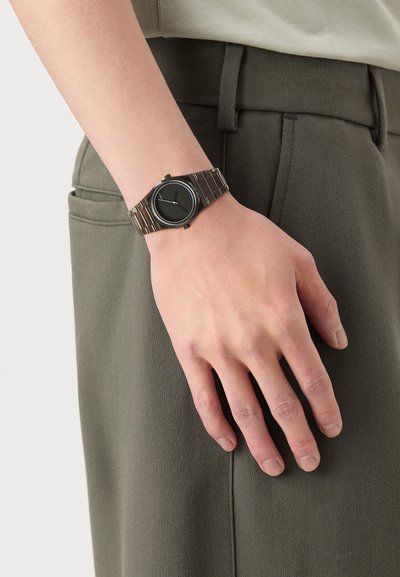 Svart klokke med rund urskive, sølv metallrem, enkle timemarkører, og gulldetaljer på kronen, båret på en hånd nær olivengrønn stoff.