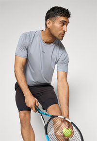 Grå idretts T-skjorte med korte ermer, med en logo, kombinert med svarte shorts. Personen holder en tennisracket og ball.
