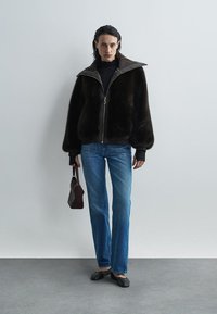 Chaqueta de pelo sintético marrón con cuello alto y cierre de cremallera; combinada con jeans rectos azules y zapatos planos negros. Sosteniendo un pequeño bolso oscuro.