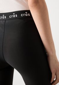 Crne tajice s glatkom teksturom i visokim strukom koje sadrže bijelo "crās" brendiranje. Bočni pregled prikazuje uski dizajn i detalje šava.