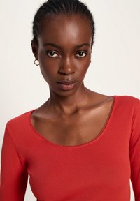 Nahaufnahme einer Frau mit geflochtenem Haar, die goldene Creolen und ein rotes Oberteil mit Rundhalsausschnitt trägt, vor einem schlichten hellen Hintergrund.