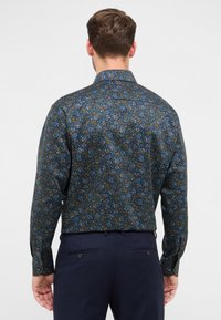 Man die met zijn rug naar de camera staat, gekleed in een donker bloemenpatroon overhemd met lange mouwen en een marineblauwe broek, tegen een effen achtergrond.