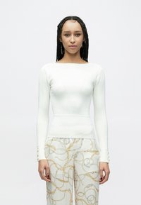 Bluză albă cu mâneci lungi și model dantelat, cu decolteu pătrat, prezentând o siluetă mulată și detalii distincte ale cusăturilor. Asortată cu pantaloni deschiși la culoare, cu model.