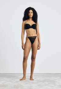 Zwart kanten lingerie set met een gepadde bh met gezaagde randen en een bijpassende slips met zilveren accenten, gedragen door een model.