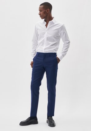 Witte button-up overhemd met een kraag en lange mouwen, gecombineerd met marineblauwe op maat gemaakte broeken. Zwarte loafers ronden de look af.