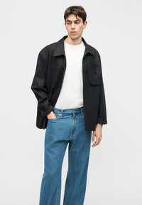 Νεαρός άνδρας που φοράει μαύρο φαρδύ μπουφάν, λευκή μπλούζα και άνετο μπλε τζιν, στέκεται με το αριστερό χέρι στην τσέπη, κοιτάζοντας προς τα πλάγια.