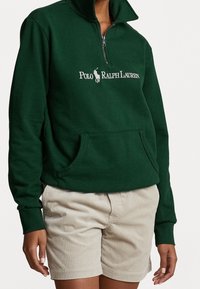 Felpa verde scuro con colletto alto e zip a metà. Presenta una tasca frontale e un logo ricamato bianco, abbinata a pantaloni corti in corduroy beige chiaro.