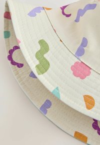 Baske i bomull i krämfärg med färgglada abstrakta former, inklusive lila, grön, orange och rosa, med broderade detaljer på brätten.
