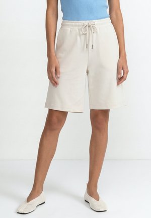 Žena nosiaca krémové kraťasy po kolená so šnúrkou, svetlomodrý tielkový top a biele pletené slip-on topánky, stojaca na bielej podlahe.