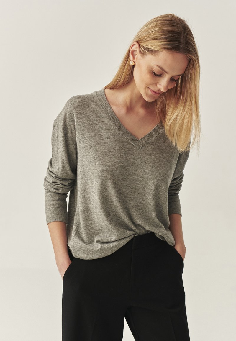 Maglione grigio a V con una vestibilità rilassata e maniche lunghe, realizzato in un tessuto morbido, abbinato a pantaloni neri. Texture liscia, design minimale.