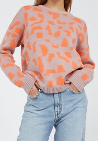 Strickpullover in zartem Rosa mit auffälligen orangefarbenen abstrakten Mustern, ausgestattet mit gerippten Bündchen und Saum. Kombiniert mit hellblauen Jeans.